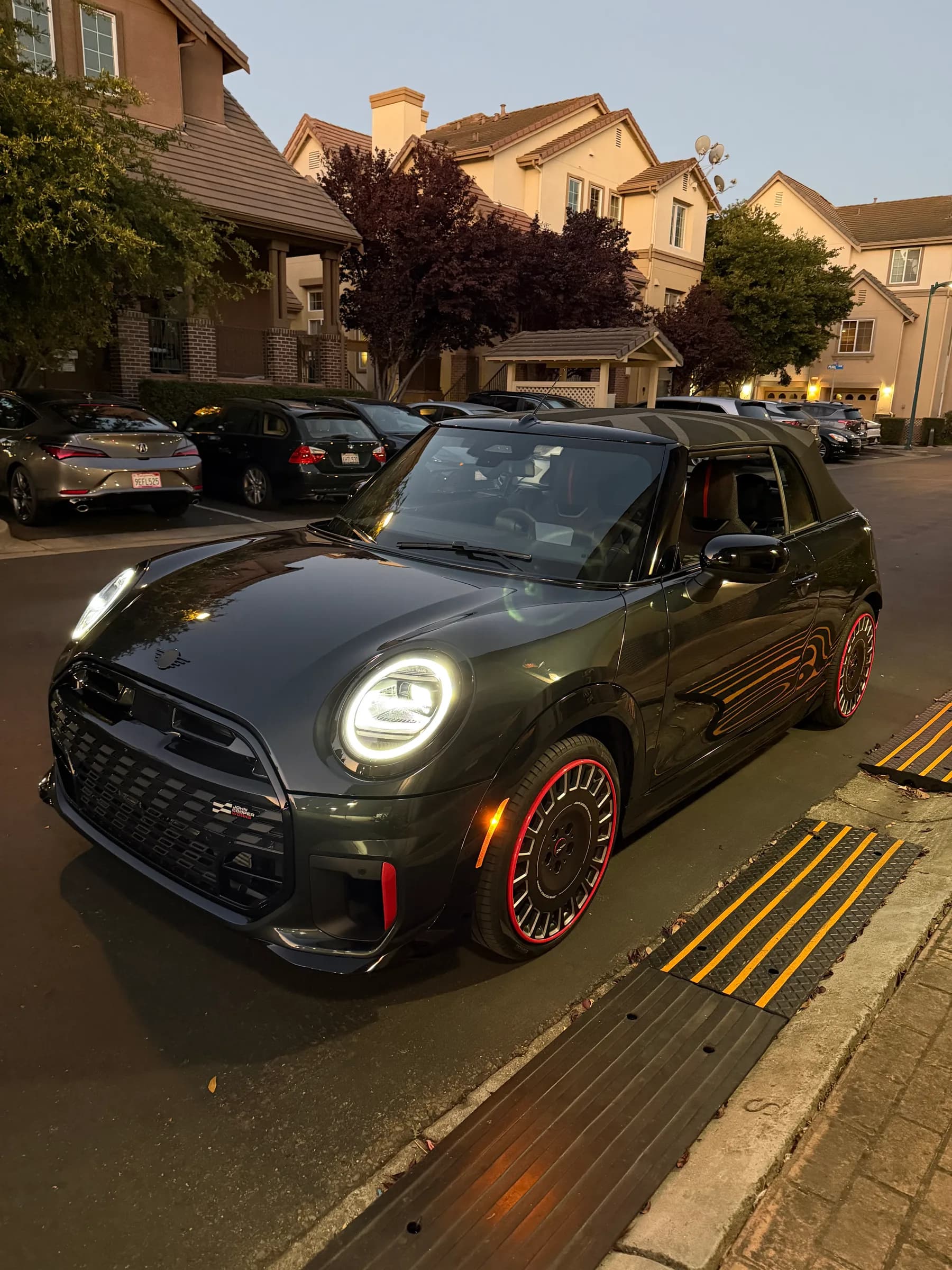 Vader, a dark gray Mini Cooper convertible with red accents, parked at a curb at night. His headlights glow, red stripes run along the side, and his dark rims a