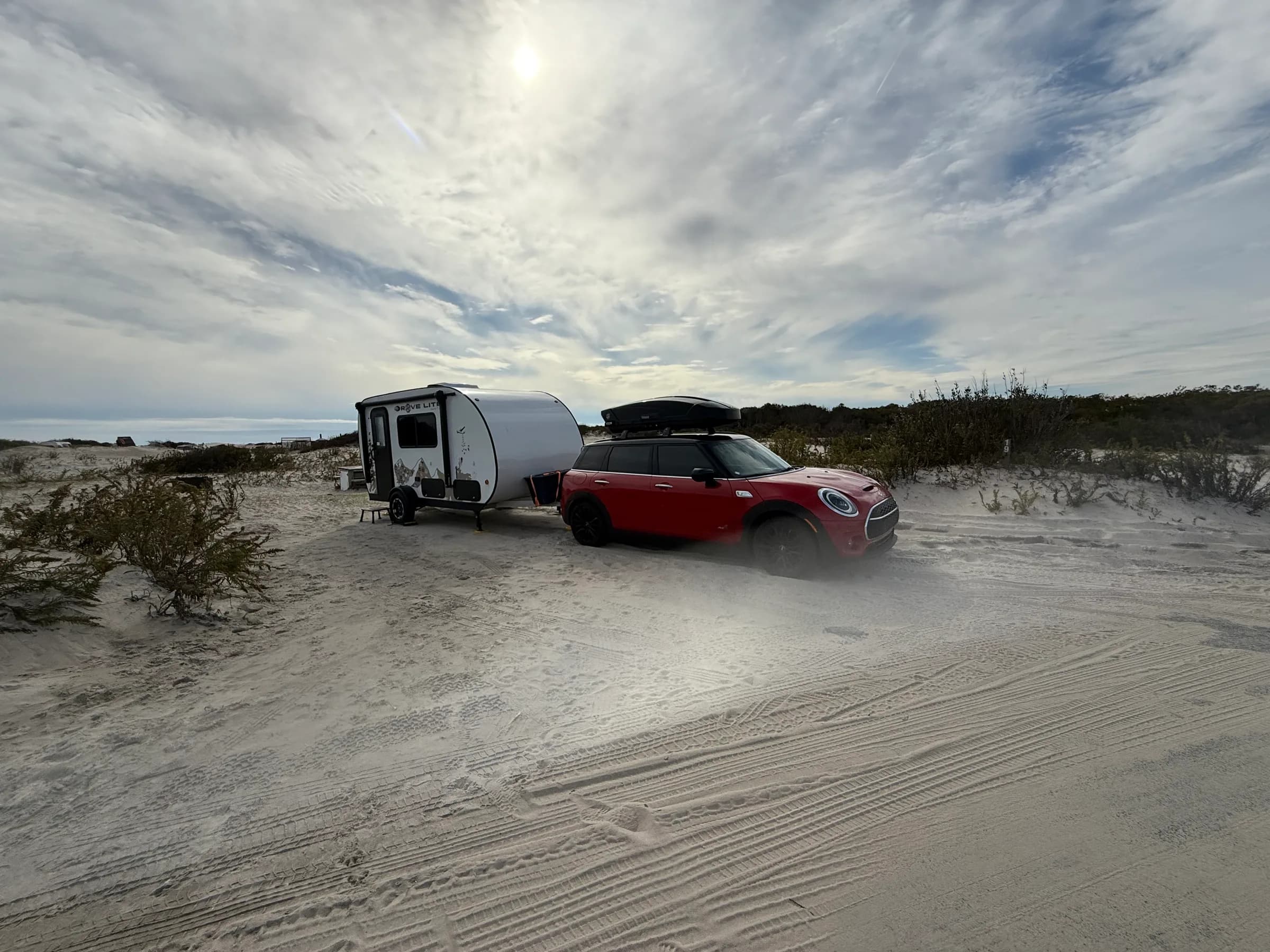 Fall camping in Assateague Park. My tiny white camper on my chilli red clubman against a raising sun, white clouds and a sandy campgrounds.